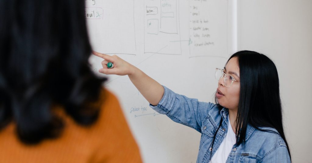 A black haired woman poiting to a whiteboard with content while another woman pays attention.