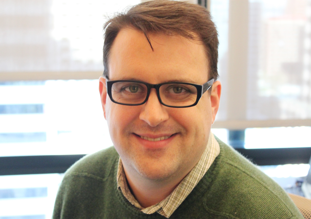 Dark brown haired man in black glasses smiling in a green sweater and a plaid shirt.