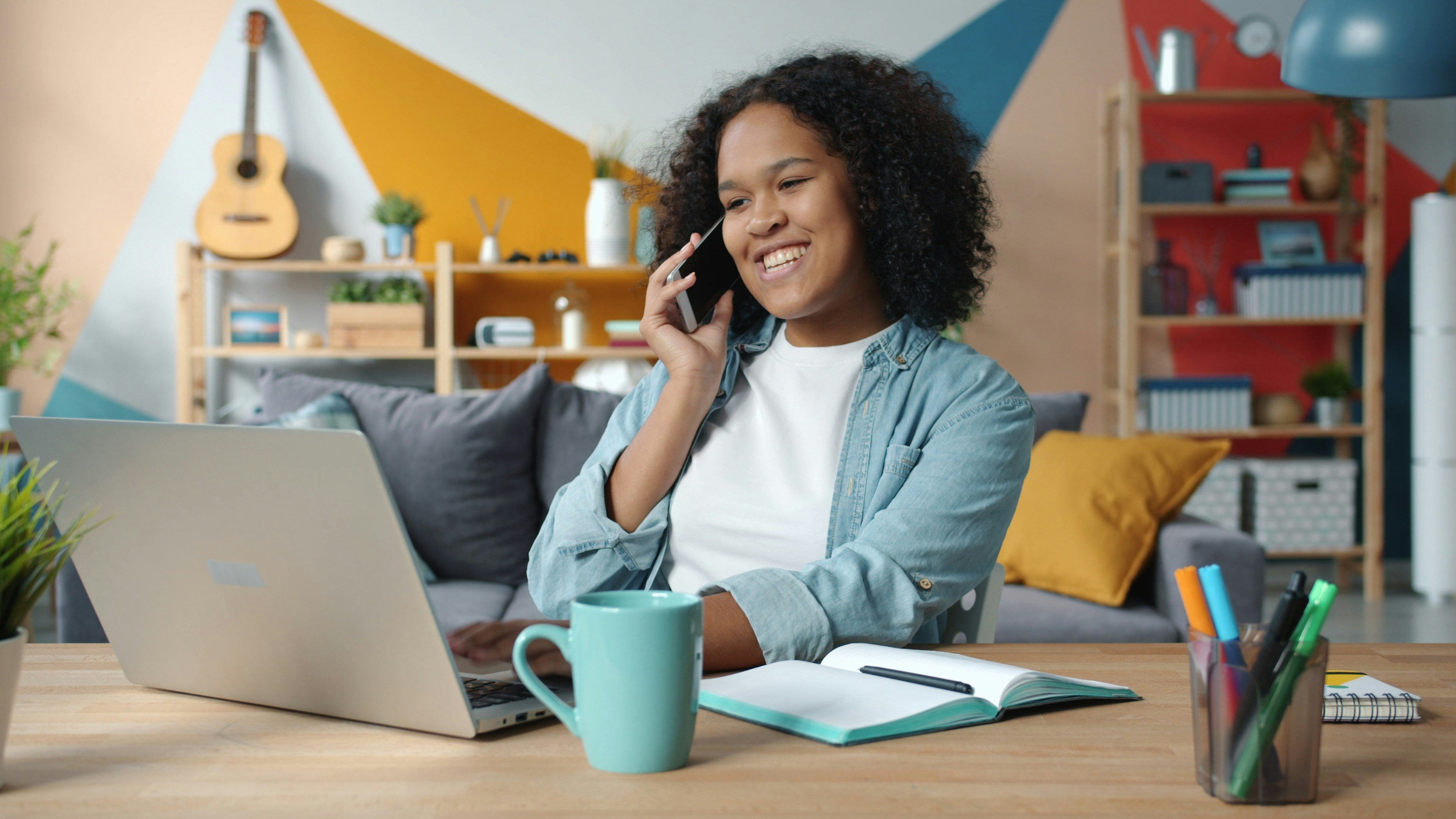 Black woman smiling, sitting at a table, talking on a mobile phone, and looking at computer screen